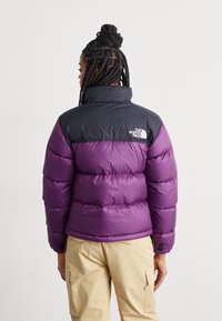 Lilla og svart pufferjakke med høy krage, med en logo på baksiden og et quiltet design. Glatt tekstur og oversized passform.
