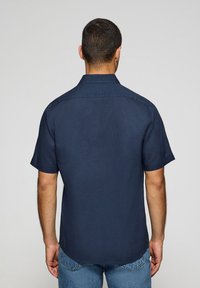 Camisa de manga corta, de color azul marino, hecha de una tela ligera con cuello normal, canesú trasero y dobladillo ligeramente curvado. Detalles mínimos.