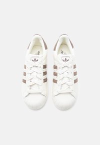 Άσπρα αθλητικά παπούτσια με τρεις καφέ σουέτ ρίγες, στρογγυλή μύτη με υφασμένο καουτσούκ, επίπεδα κορδόνια και το εμβληματικό λογότυπο στη γλώσσα.