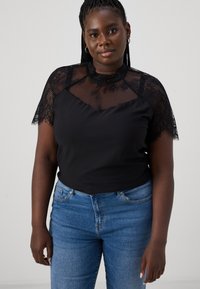 Black top with lace short sleeves, featuring a mesh panel and a rounded neckline. Paired with blue jeans for contrast.