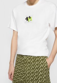 T-shirt en coton blanc présentant un motif de pièce de puzzle en noir et vert néon, assorti à un short vert olive avec un motif de texte beige.