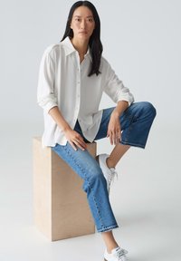 Chemise blanche à boutons avec manches longues, associée à un jean en denim bleu coupé. Le mannequin est assis sur un bloc en bois, portant des baskets blanches.