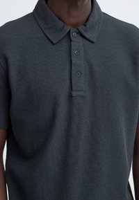 Donkergrijs poloshirt van gestructureerde gebreide stof, met een klassieke kraag en een knoopsluiting met vijf knopen. Korte mouwen, minimalistische details.