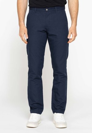 Uomo che indossa pantaloni blu scuro a gamba dritta e scarpe da ginnastica bianche, mani rilassate lungo i fianchi, in piedi davanti a uno sfondo bianco uniforme.