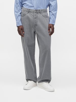 Person iført løstsiddende grå jeans, en blåstribet skjorte med knapper og hvide sneakers, der står mod en ensfarvet baggrund.
