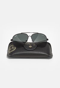 Černé sluneční brýle Ray-Ban ve stylu aviator s tmavými skly položené na texturovaném černém koženém pouzdře se zlatými logovými akcenty.