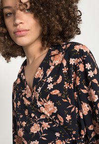 Blusa envolvente floral azul-escura com flores pêssego e brancas, mangas curtas e decote em V; textura suave com um brilho subtil.
