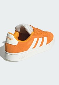 Zapatilla de Adidas de ante naranja con rayas blancas, suela blanca y cuello acolchado, vista desde la parte trasera sobre un fondo claro.