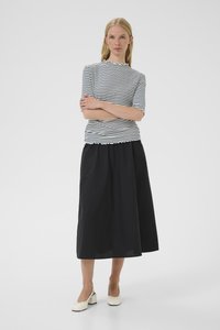 Frau mit verschränkten Armen, die einen schwarzen Midirock, ein weiß-schwarz gestreiftes Kurzarmshirt und weiße Schuhe mit Blockabsatz trägt.