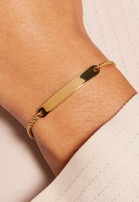 Gouden armband met een platte, rechthoekige bedel en een gestructureerd ketting; het heeft een gladde, glanzende afwerking en twee ronde bevestigingspunten.