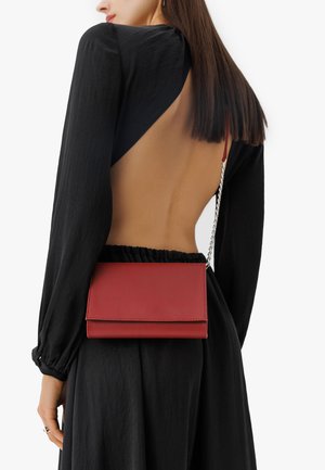 Bolso cruzado de cuero rojo con textura suave, forma rectangular y correa de cadena plateada, llevado sobre un vestido negro con la espalda al aire.