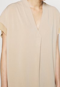 Beige kortärmad blus med V-ringning och mjukt, flödande tyg. Har en subtil struktur och minimal sömdetalj fram.