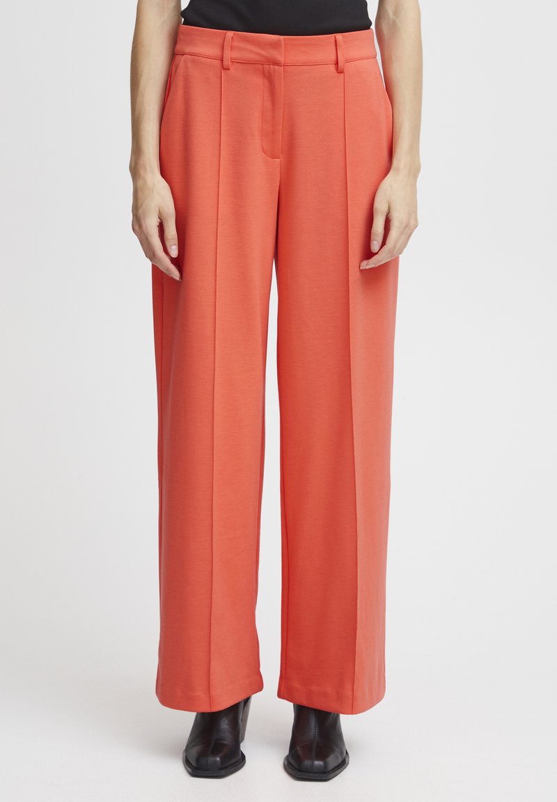 Pantalons larges orange en tissu lisse, dotés de plis à l'avant, d'une taille ajustée et de poches latérales profondes. Portés avec des bottines noires.