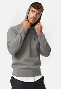 Grijze hoodie met een koord, ribgebreide manchetten en zoom. Zachte textuur; ontspannen pasvorm. Draagend over een wit shirt, trekt het model de capuchon aan.