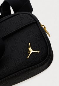 Crna torbica na rame s teksturiranim materijalom s reljefnim uzorkom "23", s zlatnim Jumpman logom kao akcentom i zatvaranjem na zip.