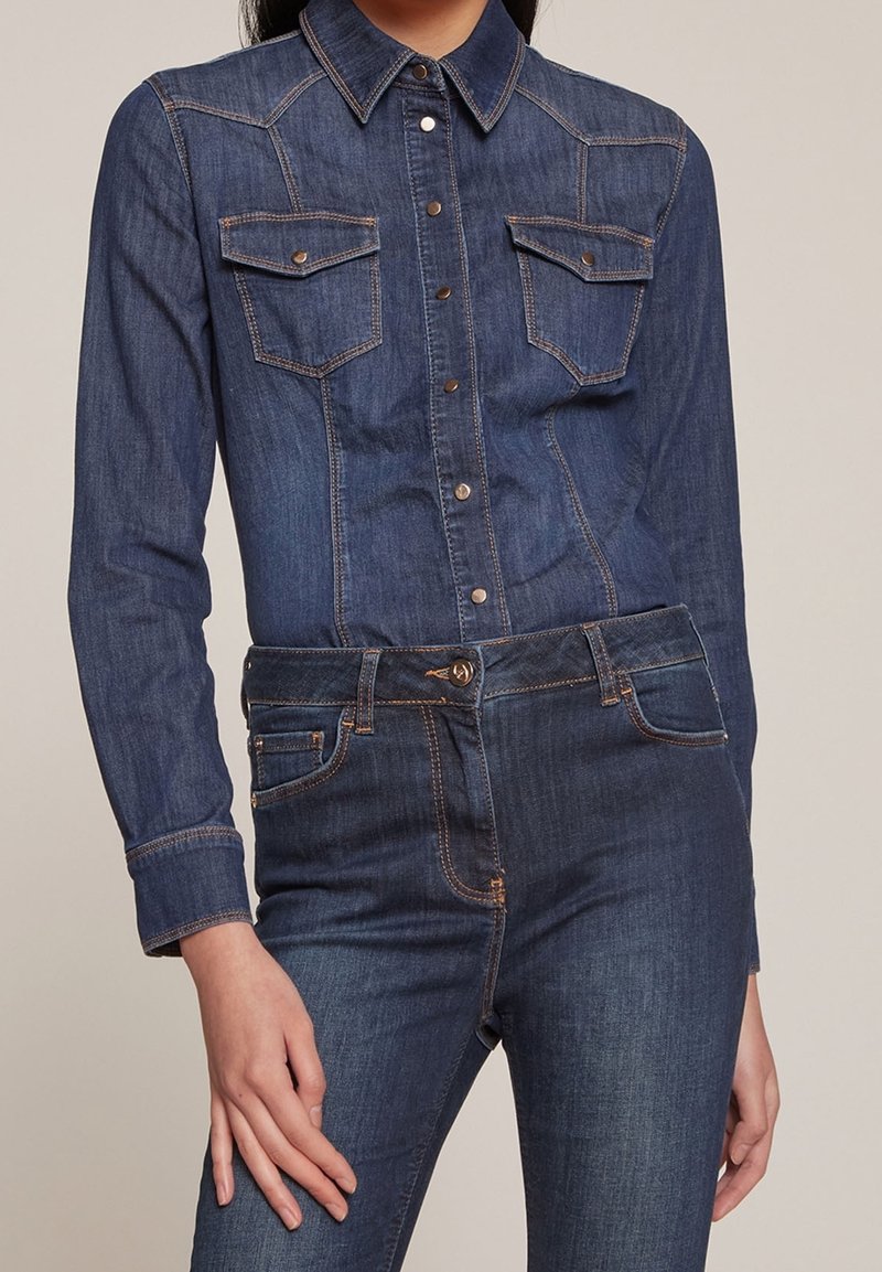 Denim shirt with long sleeves, two chest pockets, dark blue color, snap buttons, and contrasting orange stitching. Worn with matching jeans.