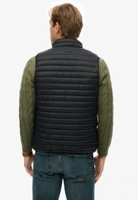 Chaleco de plumas negro con diseño acolchado, mangas en punto trenzado verde oscuro y cuello alto. Usado sobre pantalones de mezclilla azul.