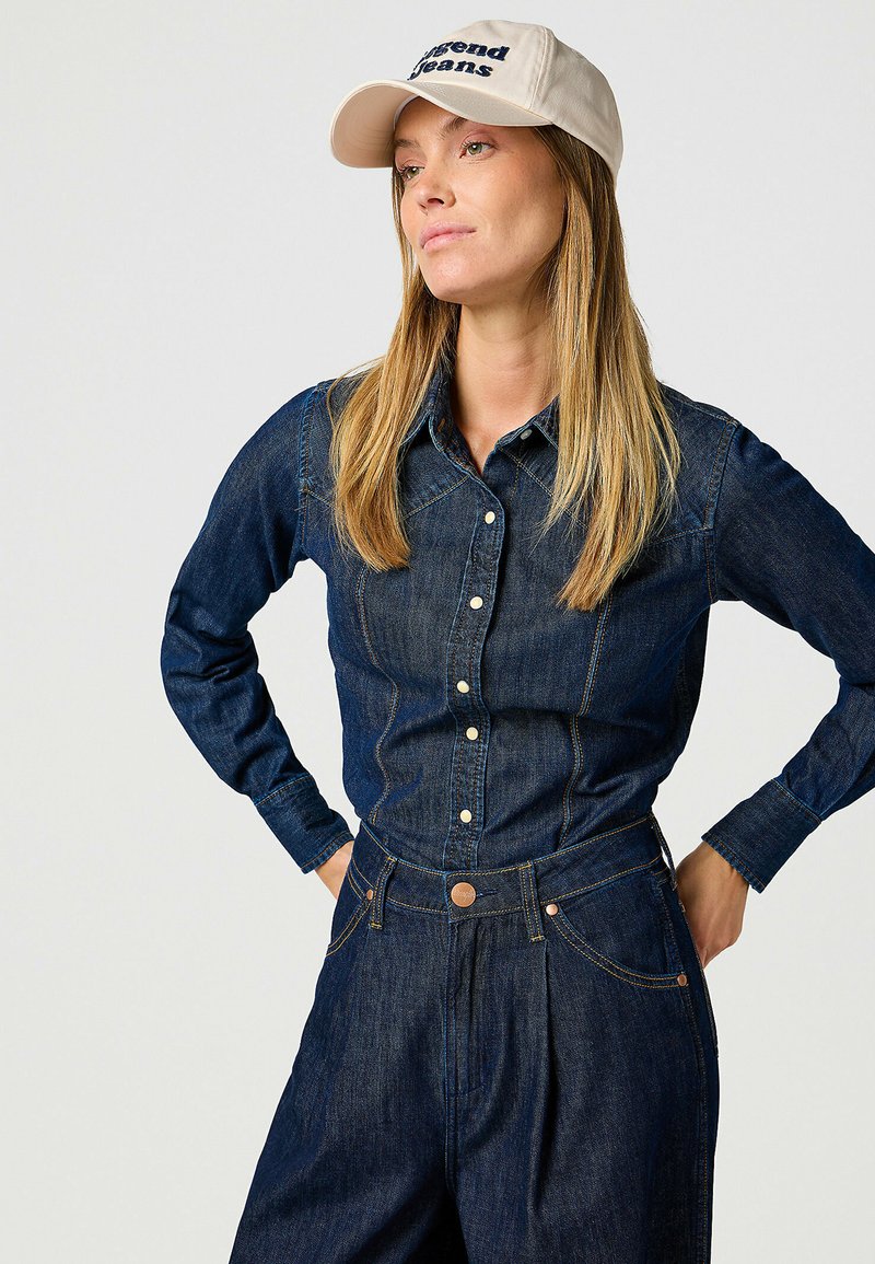 Camisa de mezclilla oscura con botón en el frente y cuello puntiagudo, combinada con pantalones de mezclilla oscuros de talle alto y una gorra beige claro con texto bordado.