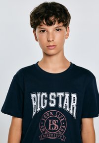 Tamsiai mėlyna medvilninė marškinėliai su balta užrašu "BIG STAR" ir apvaliu bordo logotipu, kuriame yra tekstas "TAVO GYVENIMAS NUO 1979".