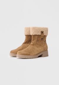 Botas de tobillo de ante en color marrón claro con un puño de borreguillo beige, que presentan una puntera redonda, suela resistente y un detalle de botón redondo en el lateral.