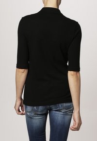 Černé polo tričko s krátkým rukávem a límečkem, vyrobené z hladkého materiálu. Přiléhavý design, pohled ze zad. V kombinaci s modrými džíny.