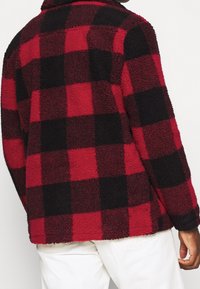 Red and black plaid jacket made of textured fleece, featuring a collar and long sleeves, with a relaxed fit and visible stitching details.