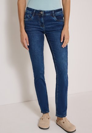 Personne portant un jean bleu ajusté, un haut bleu clair et des chaussures beiges à enfiler, debout devant un fond clair uni.