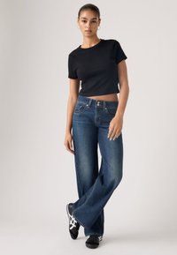 Νεαρή γυναίκα που φοράει σκούρο μπλε τζιν με φαρδιά πατζάκια, μαύρο κοντό μπλουζάκι και μαύρα αθλητικά παπούτσια με λευκές ρίγες σε απλό φόντο.