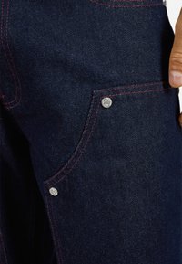 Dark blue denim pants featuring red stitching and a front pocket with a silver snap button. The fabric has a smooth texture with subtle shine.