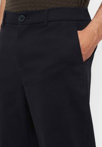 Homme portant un pantalon sombre sur mesure avec une fermeture à boutons et la main placée dans la poche avant droite.
