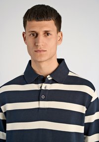 Granatowo-kremowa koszulka polo w paski z kołnierzykiem i plisą na trzy guziki, wykonana z teksturowanego materiału, z małym logo przy kołnierzyku.