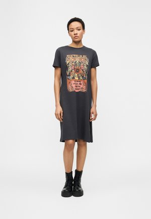 Femme portant une robe midi noire à manches courtes avec un motif floral orange et un texte, associée à des bottes noires à lacets, debout devant un fond blanc.