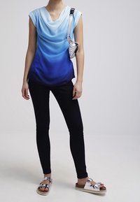 Top s kapucí v modrém gradientním ombré, spárovaný s černými skinny džínami a sandály se stříbrnými pásky. Součástí malá stříbrná crossbody taška.