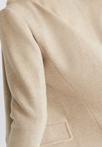 Blazer en mélange de laine beige avec une coupe ajustée, doté d'une épaule structurée, d'une poche unique et d'une finition lisse et texturée.