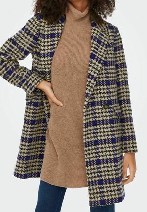 Kvinde iført blå og beige hundetandsmønstret frakke over en beige strikket coltrøjekjole med blå jeans, hånd på hoften, mod en hvid baggrund.