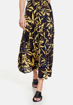 Femme portant une jupe midi à motifs jaune, marine et marron avec des mules à talons ouvertes noires, marchant sur un sol blanc.