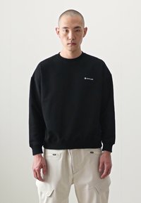 Sweatshirt noir à col ras du cou avec logo sur la poitrine gauche, associé à un pantalon cargo blanc avec poches avant et un tissu texturé.