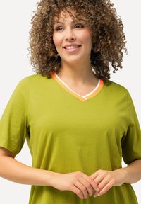 Zelené tričko s krátkým rukávem a výstřihem do V, s hladkým povrchem, zdobené oranžovým a bílým lemem na límci. Model si drží ruce pohromadě.