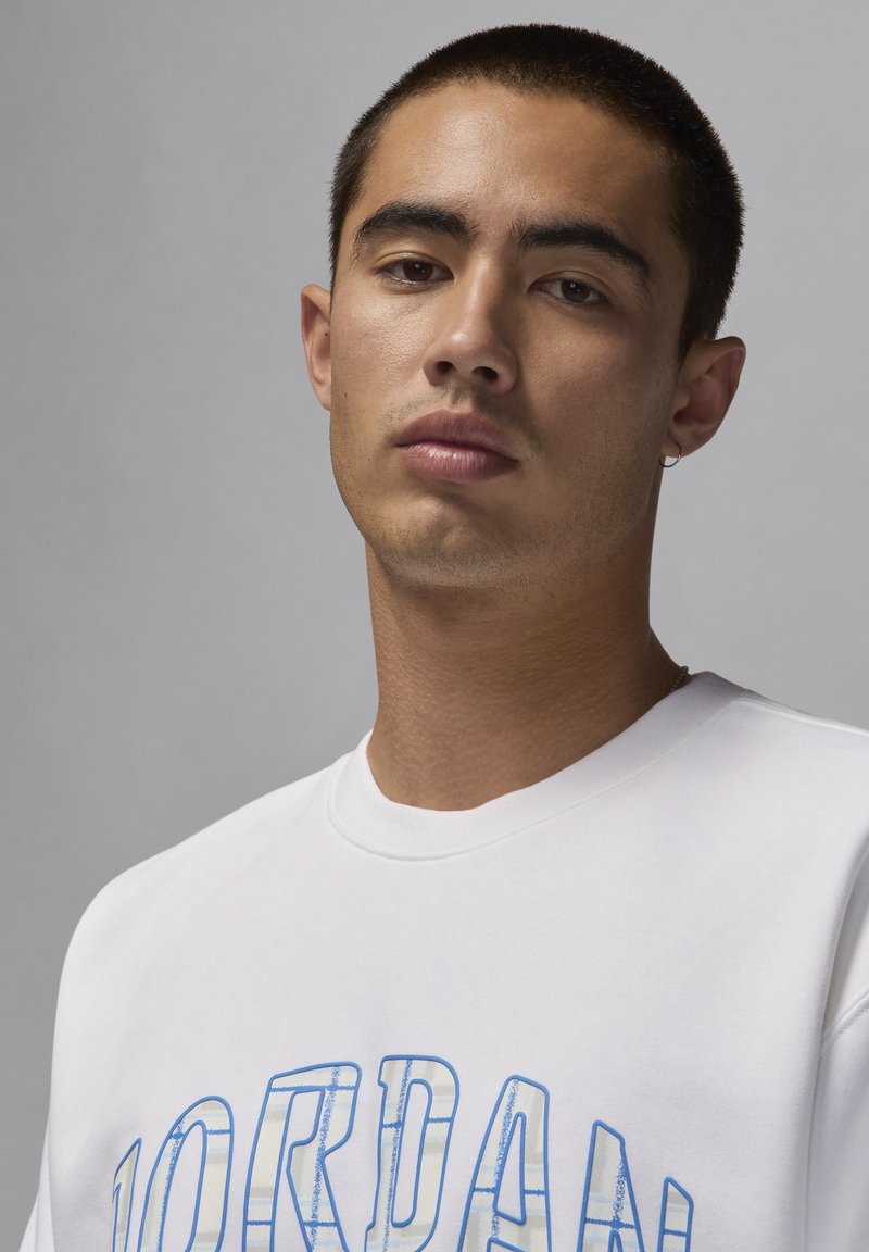 Sweatshirt blanc avec graphique « JORDAN » bleu. Matière en coton lisse, col rond et manches courtes. Modèle avec cheveux courts et boucle d'oreille.