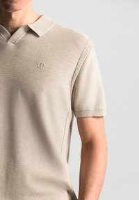 Beige polo krekls, izgatavots no teksturēta auduma, ar V veida kaklu, īsām piedurknēm un izšūtu logo uz kreisā krūts.
