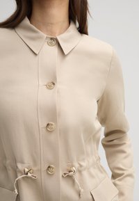 Giacca button-up beige chiaro con colletto ampio, vita regolabile con lacci e due tasche frontali; presenta bottoni crema e dettagli in hardware dorato.