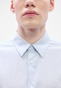 Camisa de vestir de rayas azul claro con cuello clásico, dos botones en el cierre y un logo bordado en el pecho. La tela parece suave.