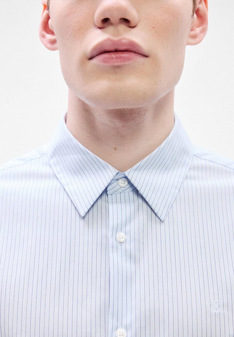 Hellblau gestreiftes Hemd mit klassischem Kragen, zwei Knopfverschlüssen und einem aufgestickten Logo auf der Brust. Der Stoff wirkt glatt.