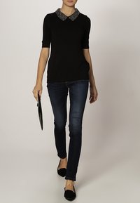 Kvinna som går klädd i en svart kortärmad topp med en glittrig krage, mörka smala jeans, svarta platta skor och håller en liten svart kuvertväska.