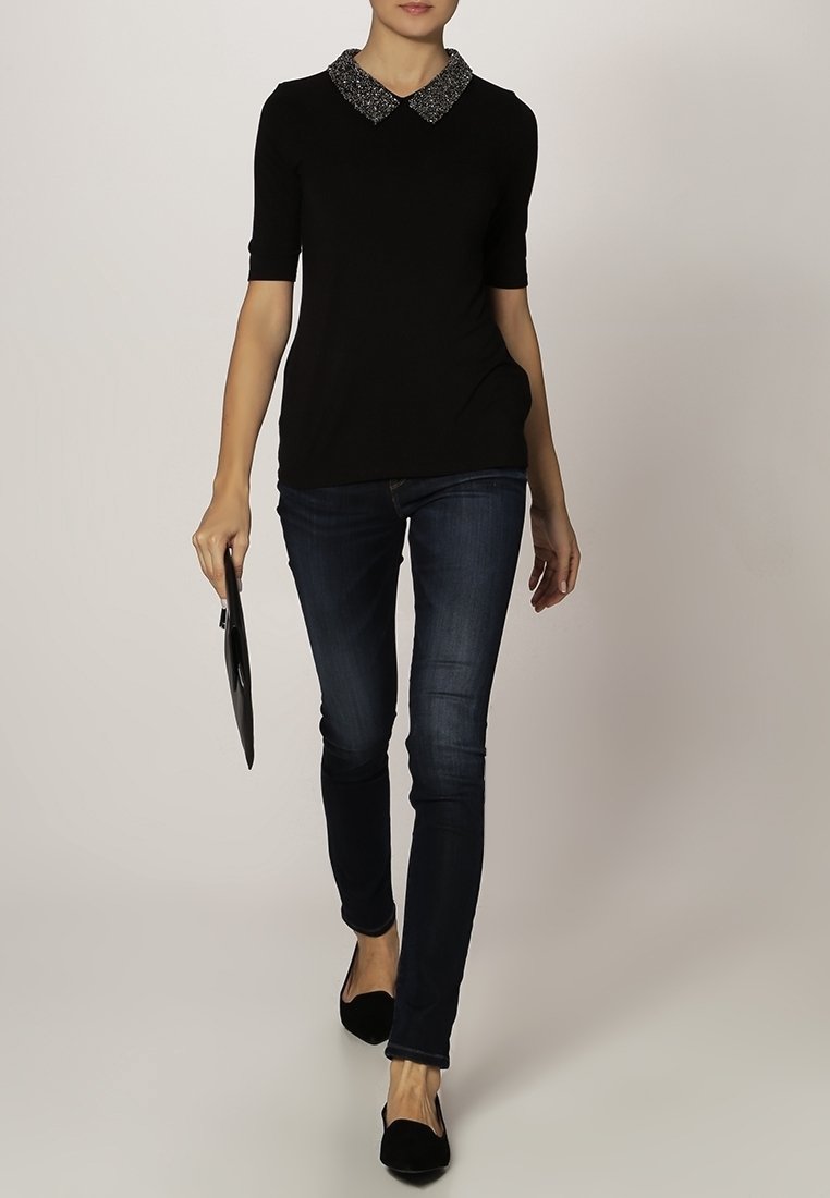 Kvinna som går klädd i en svart kortärmad topp med en glittrig krage, mörka smala jeans, svarta platta skor och håller en liten svart kuvertväska.