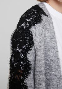 Grijze gebreide cardigan met zwarte kanten details op één mouw. Kenmerkt zich door een zachte textuur en een losse pasvorm, die het contrast tussen de materialen benadrukt.