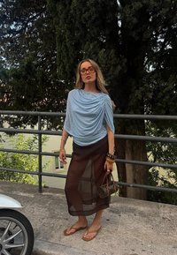 Haut drapé bleu clair avec des embellissements, jupe brune transparente, flip-flops bruns et un petit sac à main à motif. Foliage vert en arrière-plan.