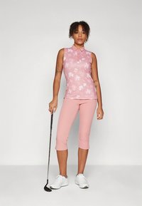 Πόλο μπλούζα σε ανοιχτό ροζ με λουλουδάτο μοτίβο, συνδυασμένη με ανοιχτό ροζ κάπρι παντελόνια. Το μοντέλο κρατάει ένα γκολφ κλαμπ και φοράει λευκά αθλητικά παπούτσια.