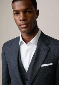 Blazer testurizzato blu scuro con rever a punta, indossato sopra una camicia bianca. Presenta un fazzoletto bianco nella tasca e dettagli in bottone nero.