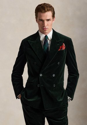 Homme portant un costume croisé en velours vert foncé avec cravate, chemise blanche, pochette rouge, mains dans les poches et expression sérieuse.